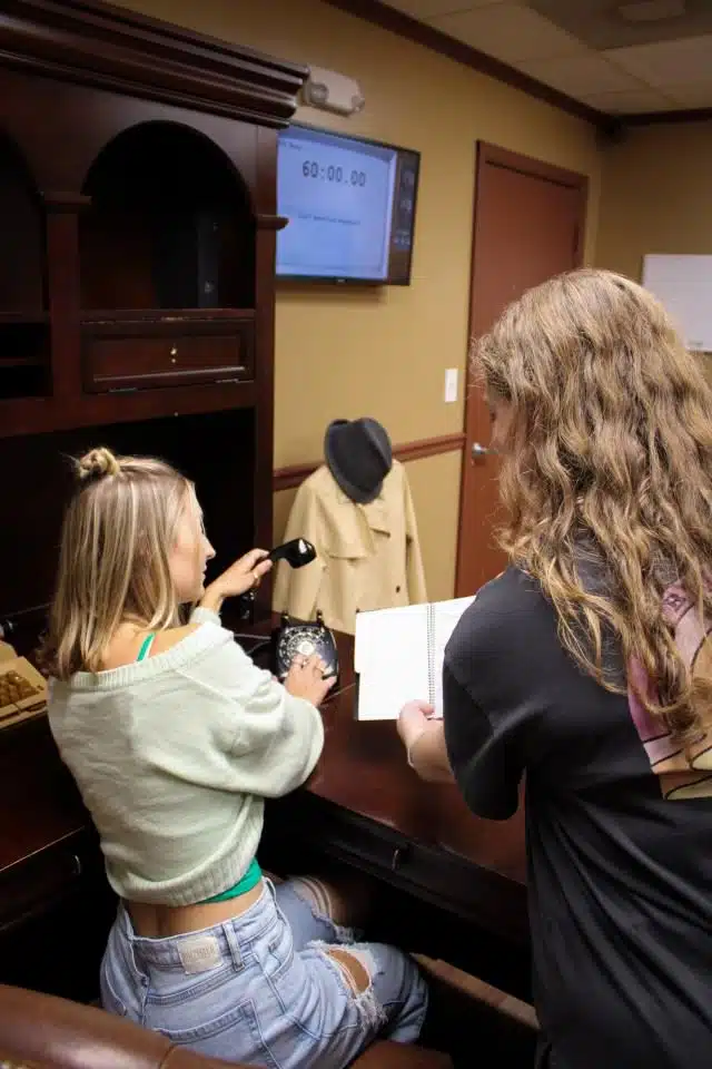 An image of the Mob Boss at Escape This Frederick. Two girls are seen looking into a book and picking up a rotary phone.