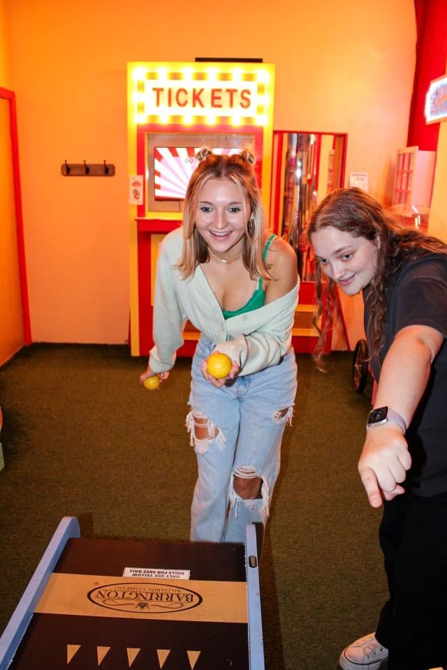 An image of The Carnival escape room at Escape This Frederick. Two players are seem smiling while one girl throws Skee Balls and the other points towards the game.