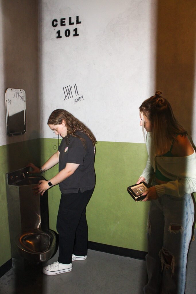 Image of the Cell Block 1 escape room. Two girls inspecting a sink and a badge.